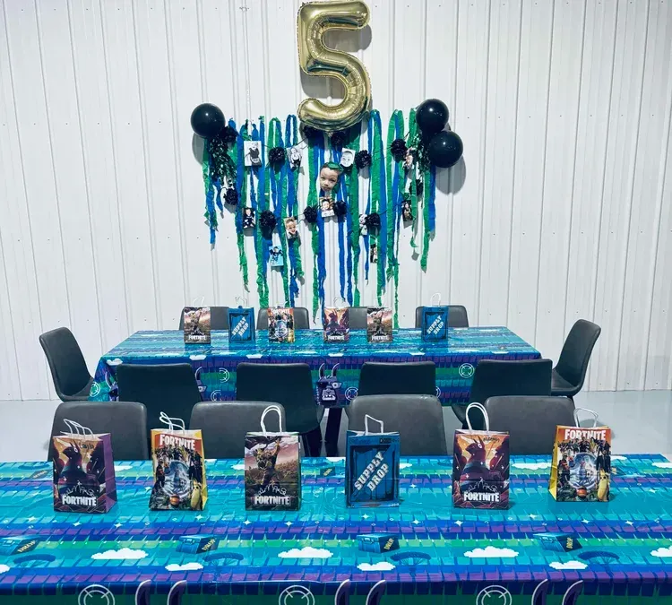 Fortnite-themed birthday table: blue and green decorations, party favors, number 5 balloon, black chairs.