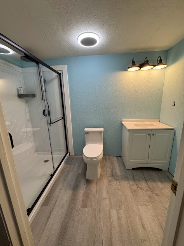 A bright, renovated bathroom featuring a glass shower, toilet, and white vanity against light blue walls and wood floors.