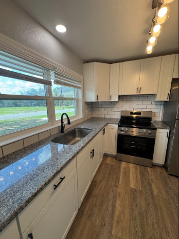 A kitchen with white cabinets, granite countertops, a stainless steel oven, a sink by a window, and wood-look flooring.