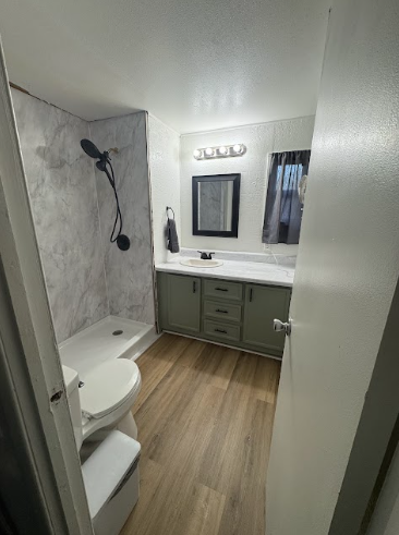 A small, renovated bathroom with light wood flooring, sage green vanity cabinets, a white shower, and a toilet.