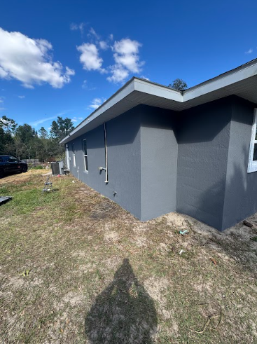 A corner exterior view of a grey stucco house under a bright blue sky with light clouds.