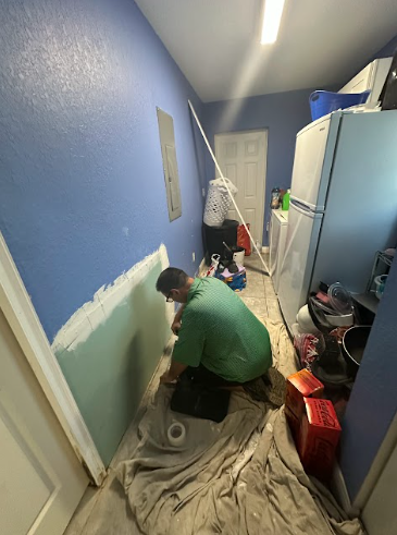 A person in a green shirt kneels on a drop cloth in a blue laundry room, applying joint compound to a patch of drywall.