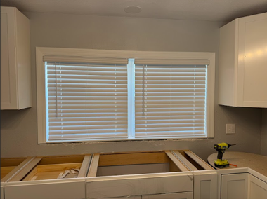 White kitchen cabinets frame a window with closed blinds above unfinished base cabinets, with a drill on the counter.