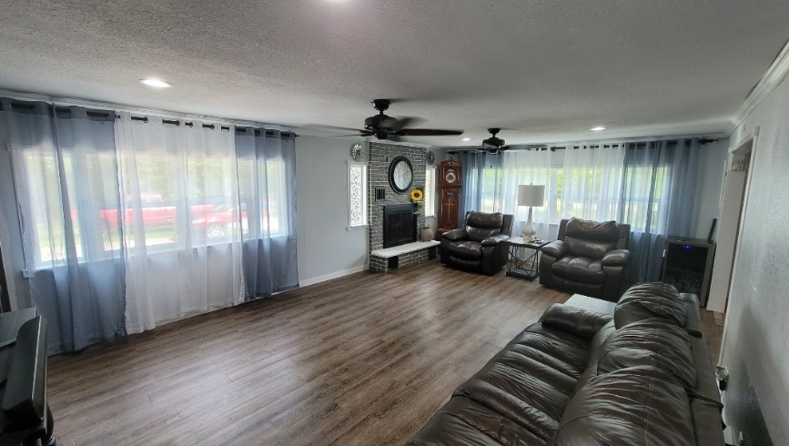 A spacious living room with wood flooring, large windows with layered blue and white curtains, a fireplace, and seating.