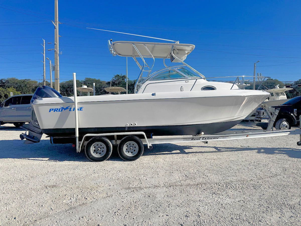 White boat on a trailer, parked on gravel with blue sky in background. 