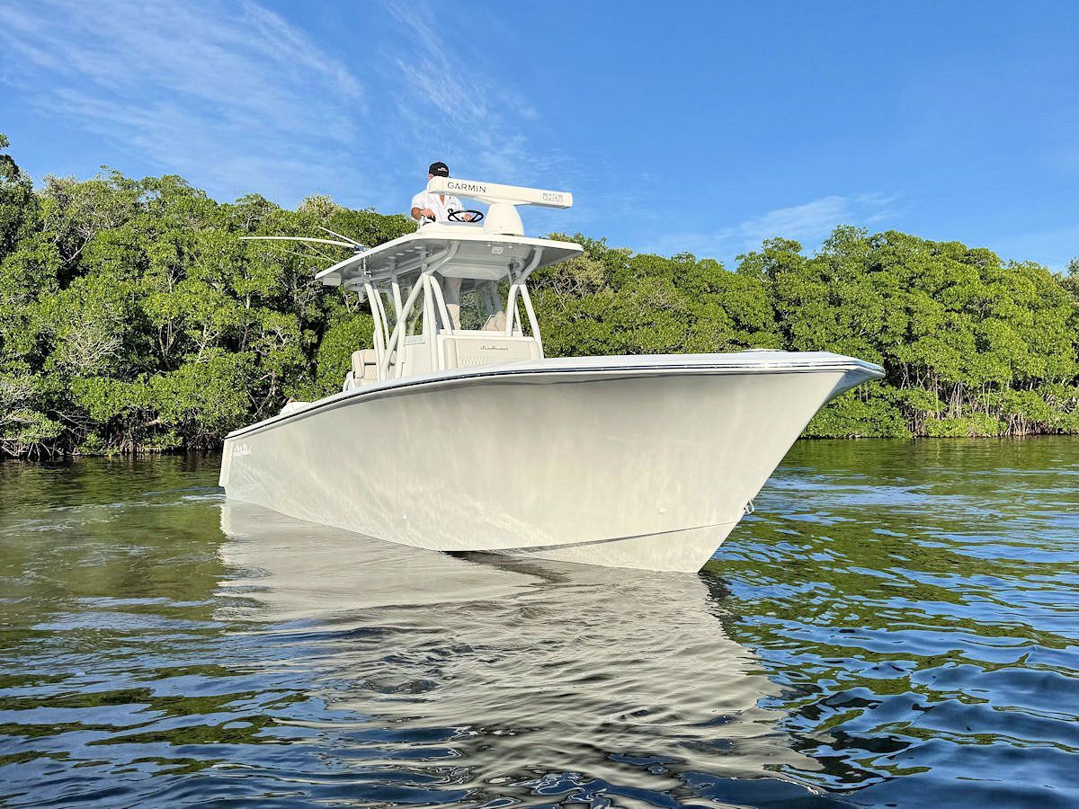White motorboat on water, person at the helm, trees in background, sunny day.