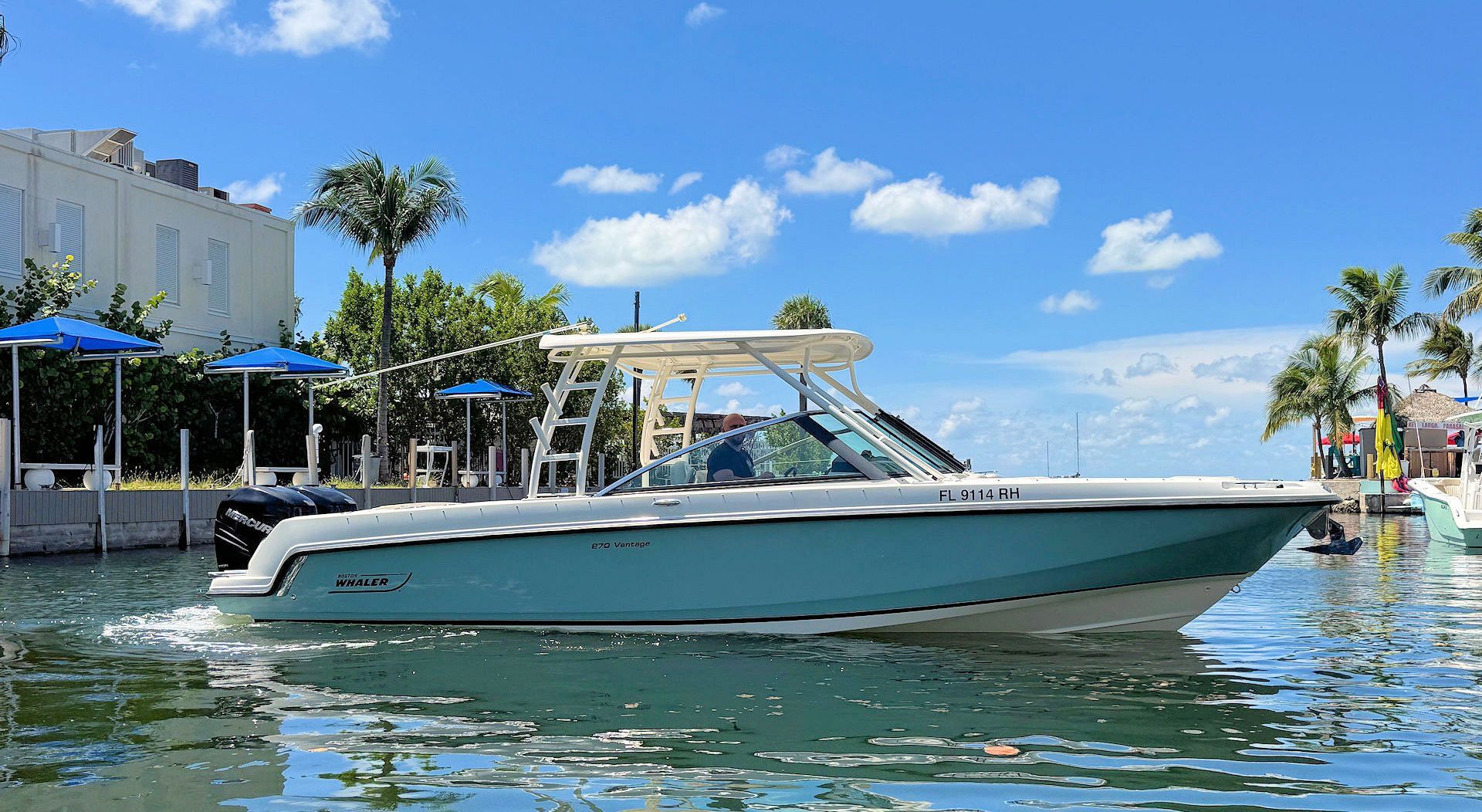 A teal and white motorboat moves across calm water near a marina with palm trees under a bright blue, sunny sky.