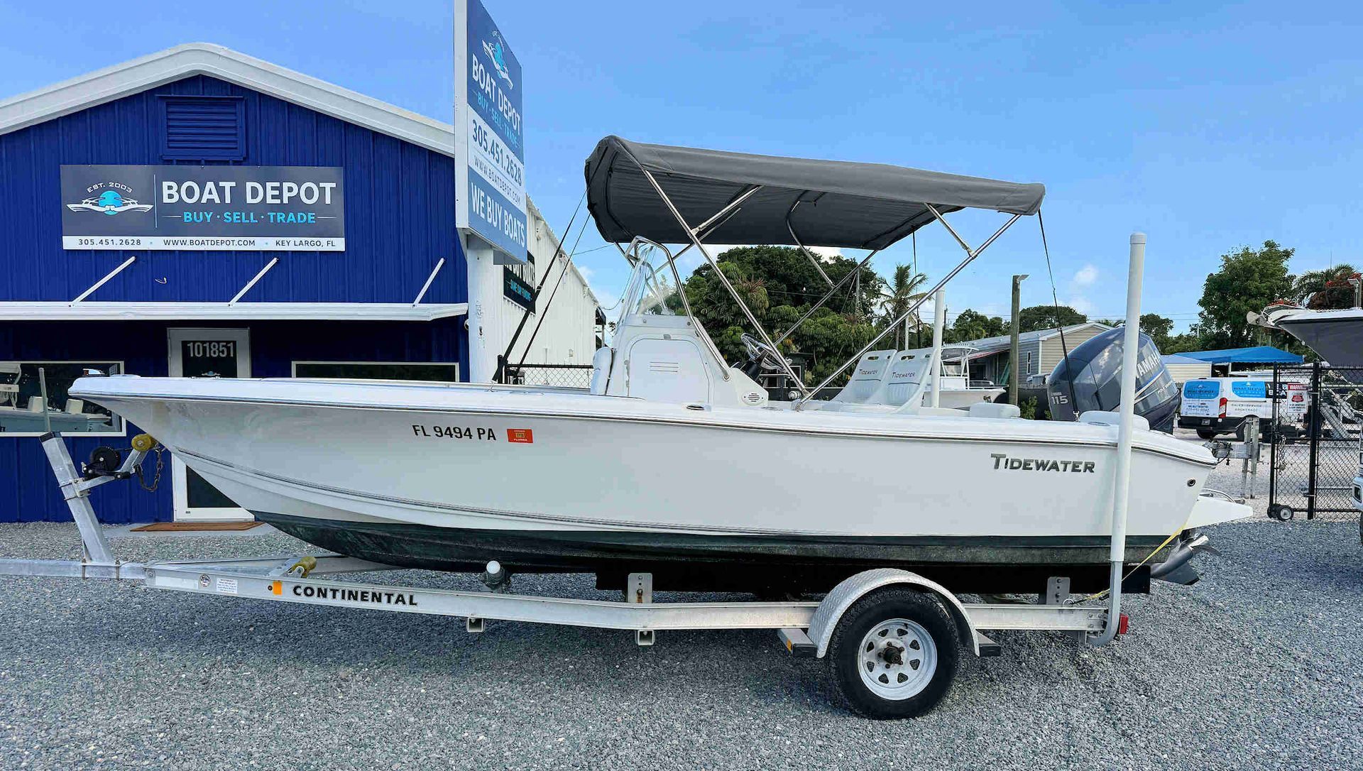 White Tidewater skiff on a trailer outside a blue boat dealership building on gravel, under a clear sky