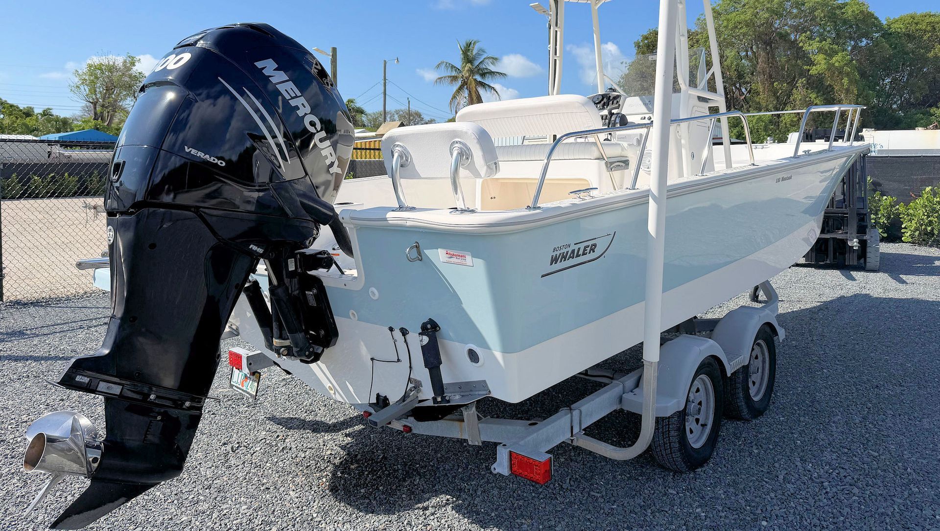 White boat with black outboard motor on a trailer, parked on gravel under a sunny blue sky.