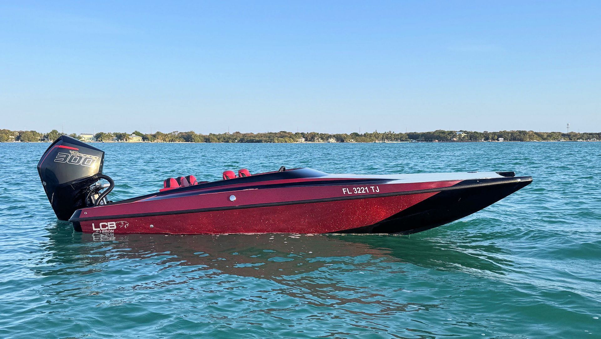 Red high-performance catamaran on calm blue water with a Mercury 330R, near a low shoreline under a clear sky