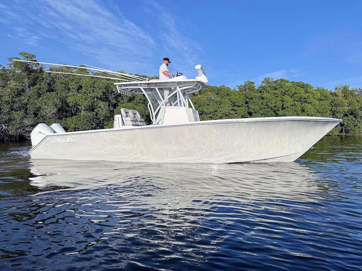 White motorboat on water with a person at the helm, trees in the background, sunny day.