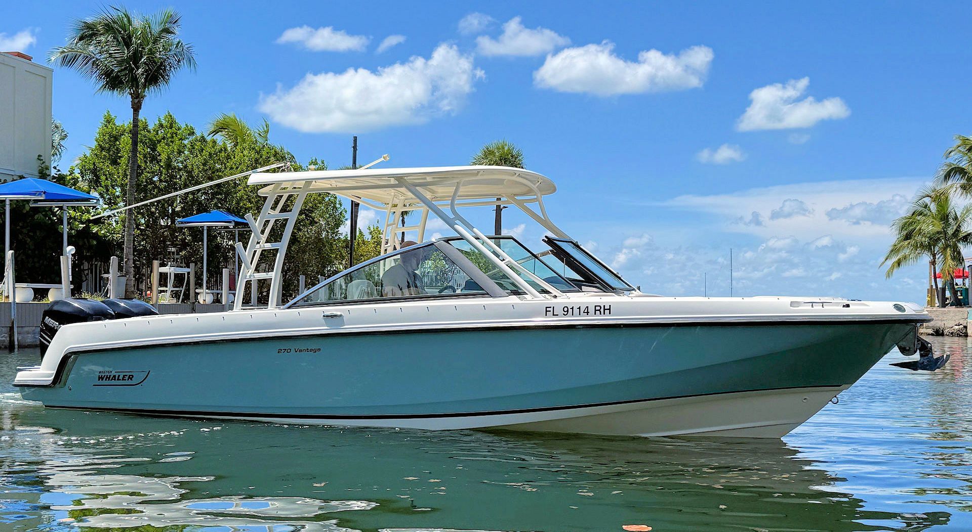 A light blue Boston Whaler motorboat with a white hardtop cruising on sunny tropical waters near a dock.