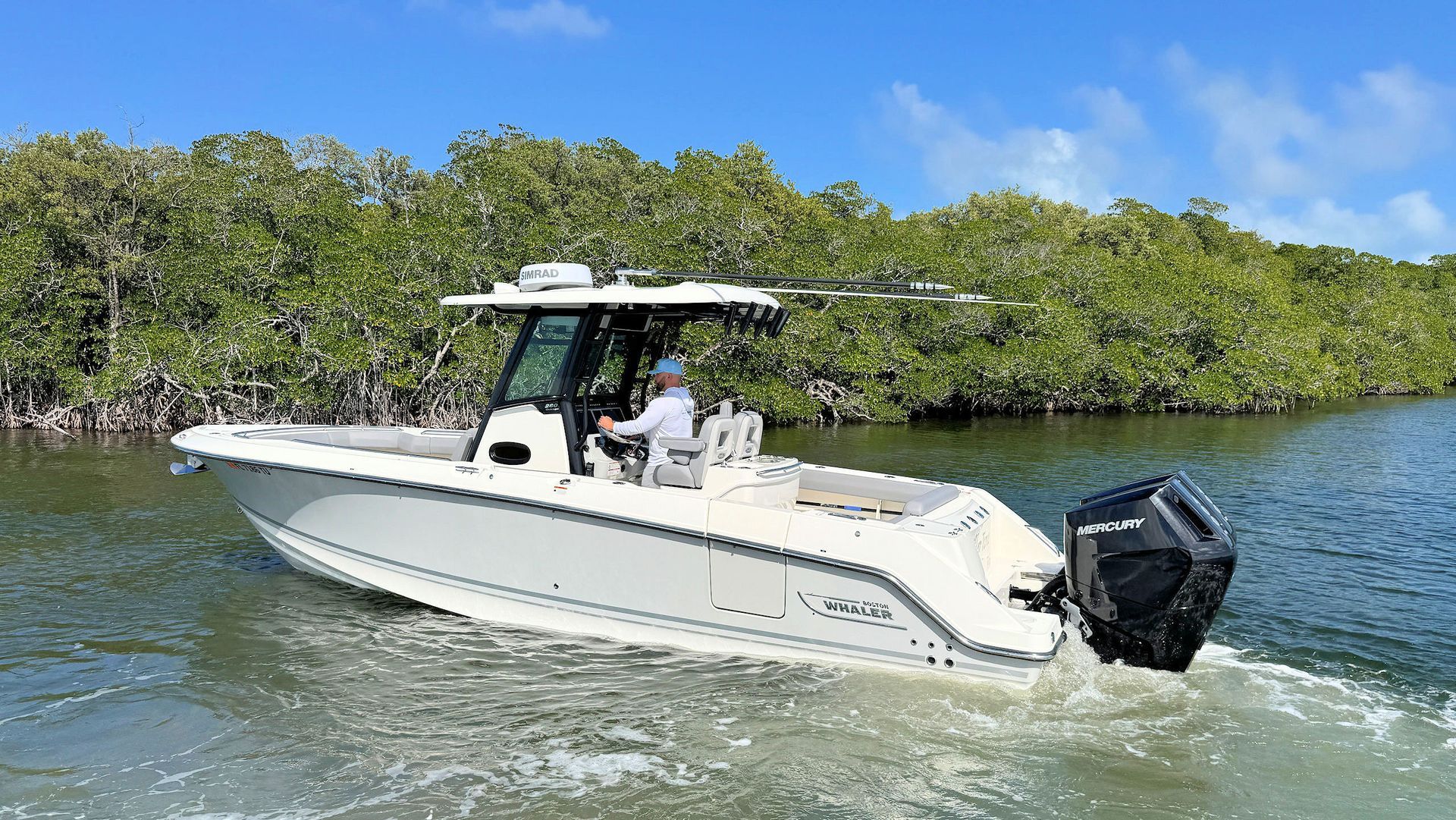 A person steers a white Boston Whaler center console motorboat through calm water near a lush mangrove shoreline.