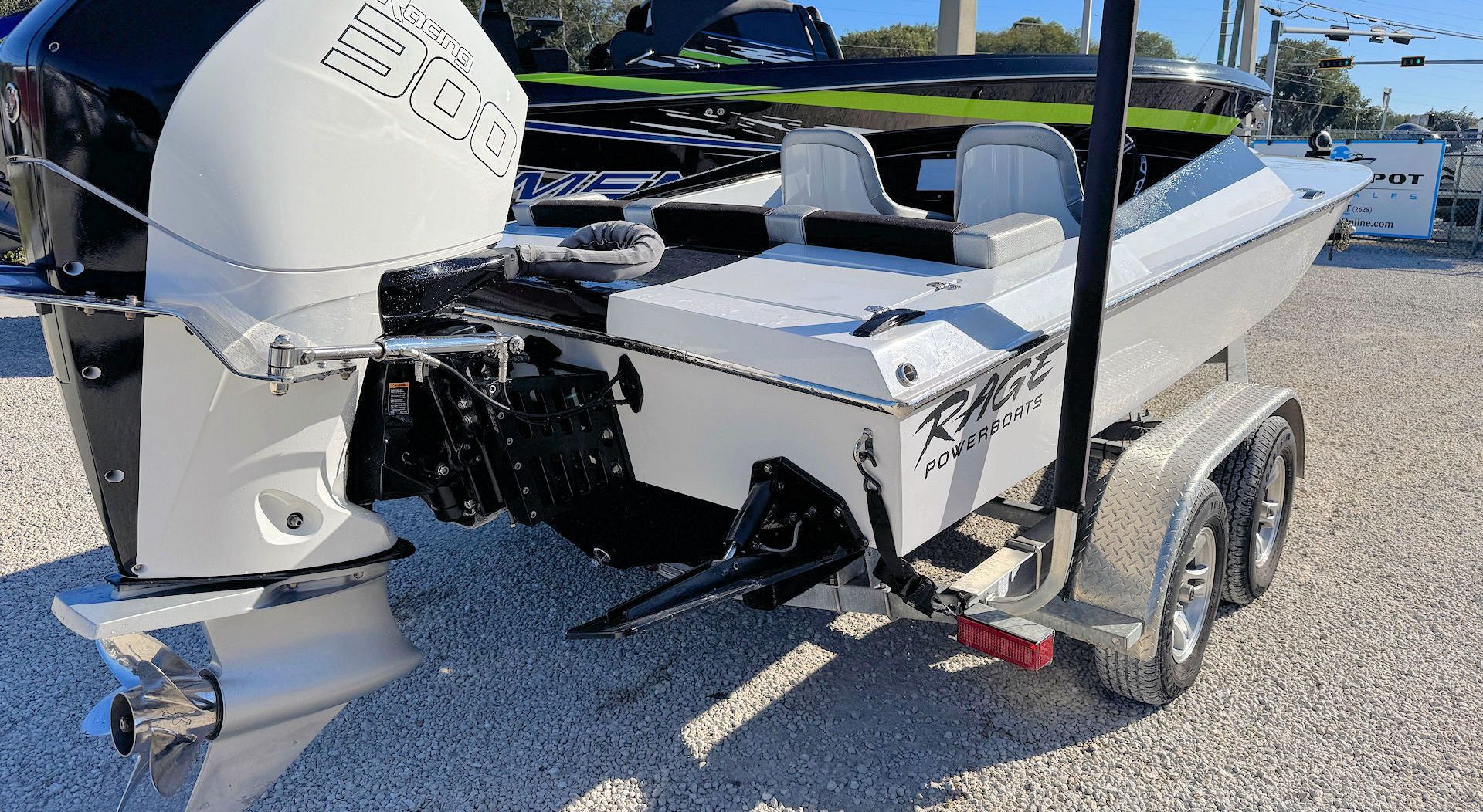 White motorboat with a black outboard motor on a trailer, parked on a gravel lot under a clear blue sky.