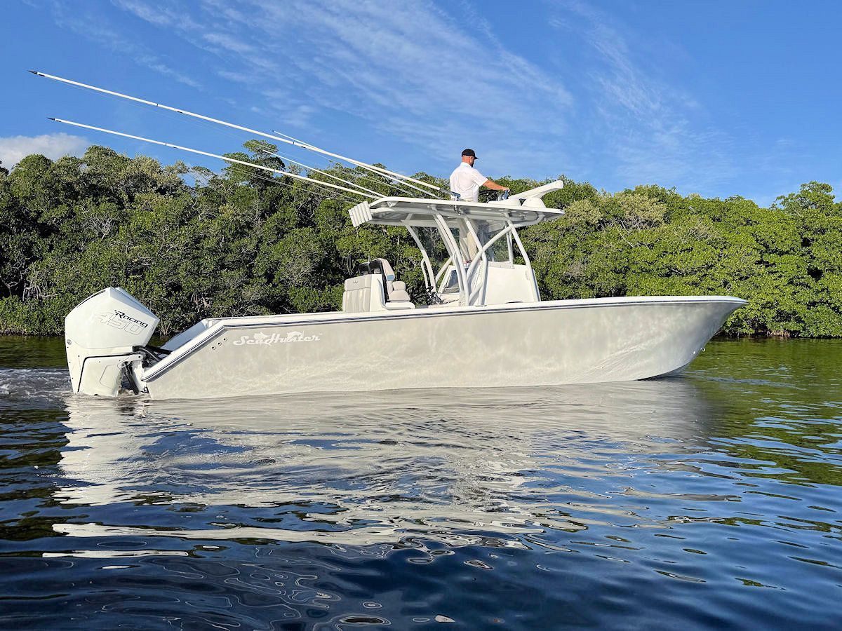 A man stands on a silver boat in the water, near green trees under a blue sky.
