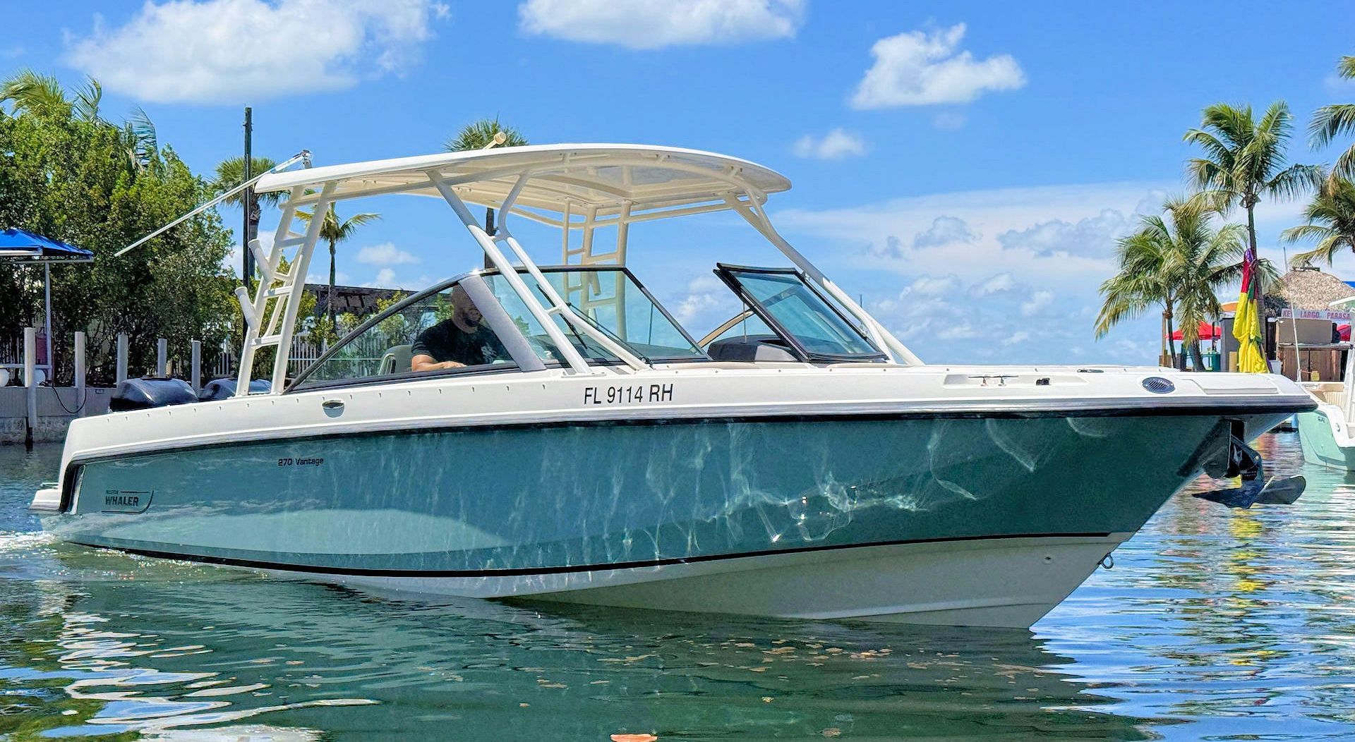 A teal-hulled dual-console boat with a hardtop navigates calm blue tropical waters near a shoreline with palm trees.