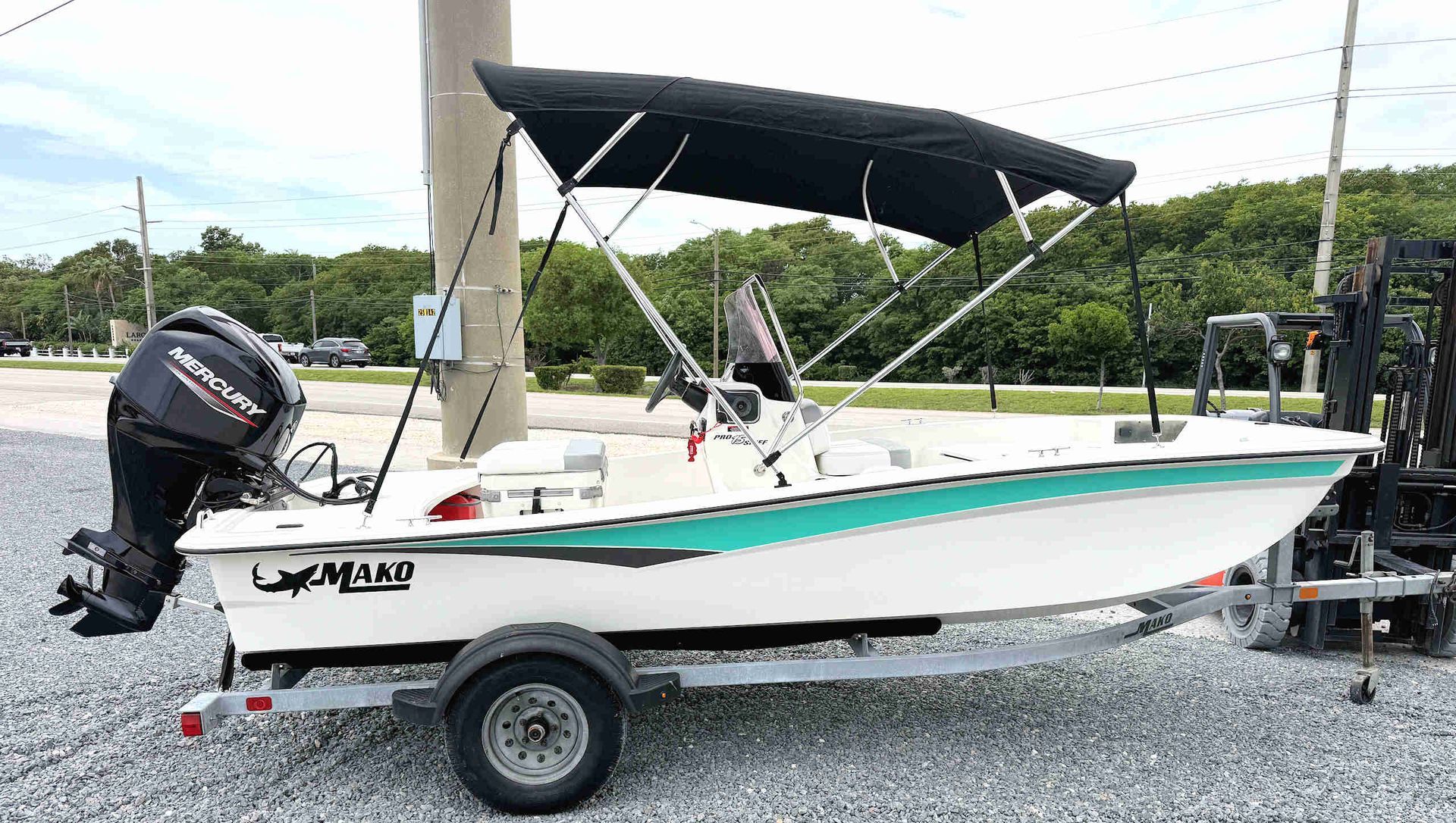 White fishing boat on a trailer with black bimini top and black outboard motor, parked on gravel.