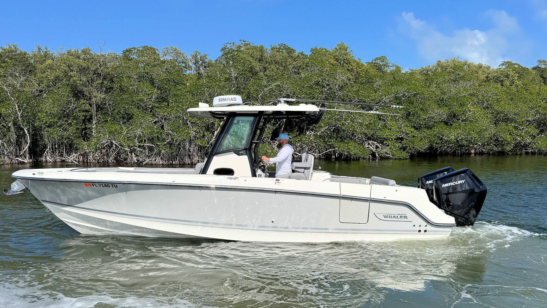 A white Boston Whaler center-console boat cruises through calm water near a shoreline lined with lush green mangrove trees.