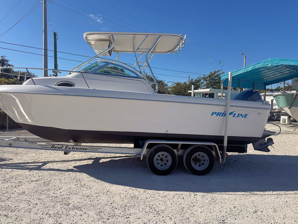 White Pro-Line boat on a trailer, parked on gravel with a sunny sky and other boats nearby.