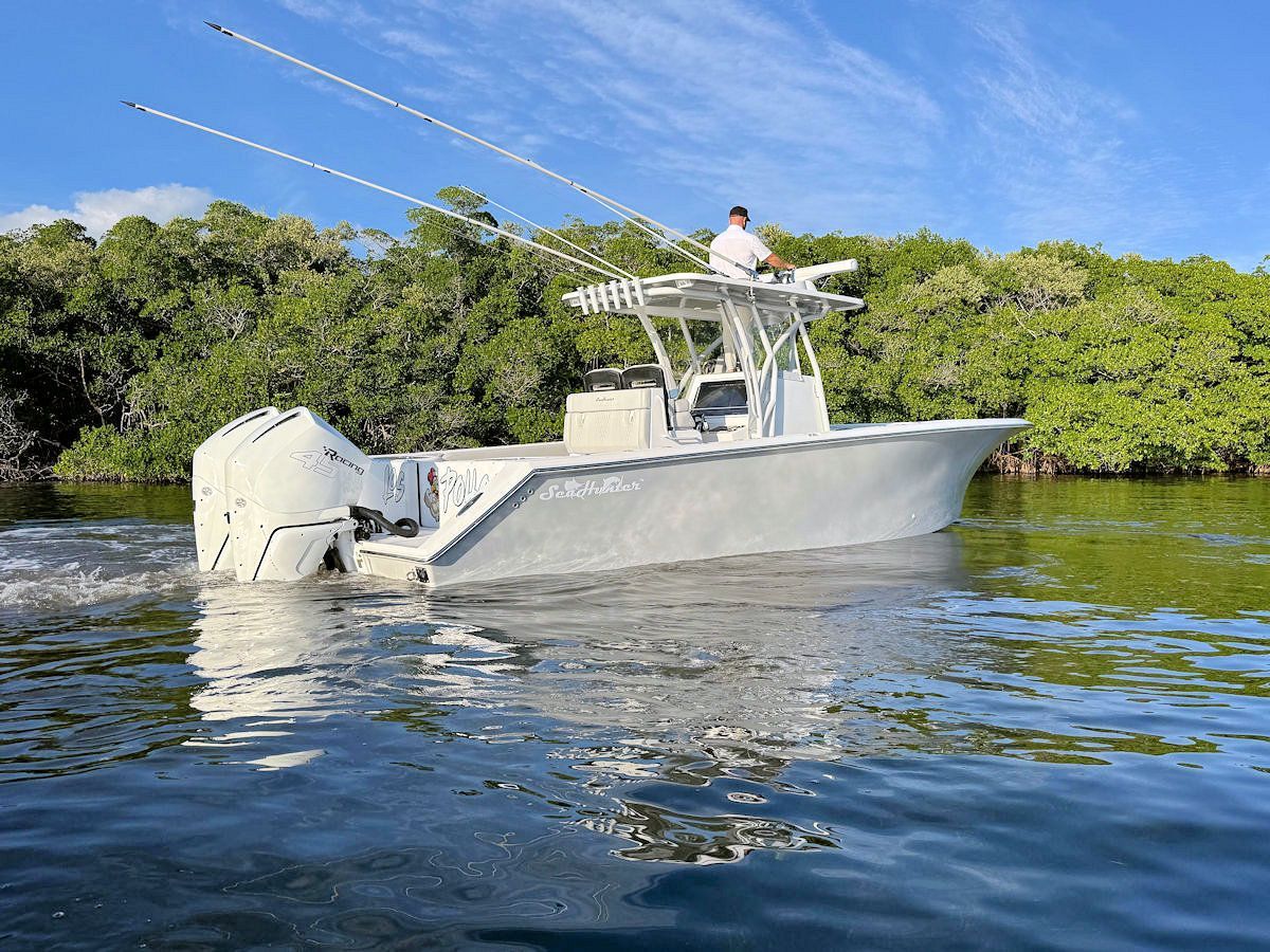 A white fishing boat with a person at the helm on water near green foliage.