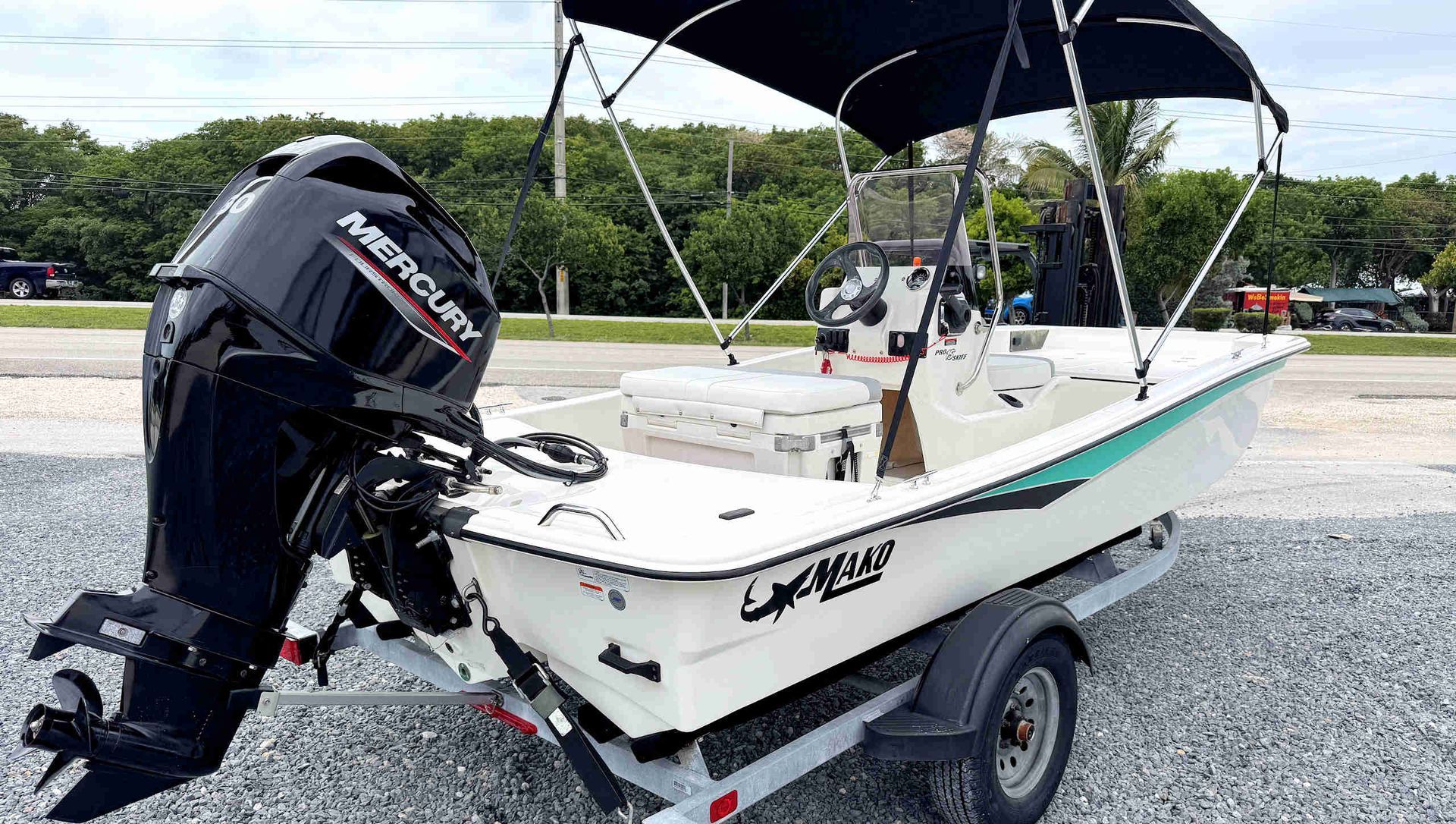 Small white fishing boat on a trailer with black outboard motor and canopy in a gravel lot.