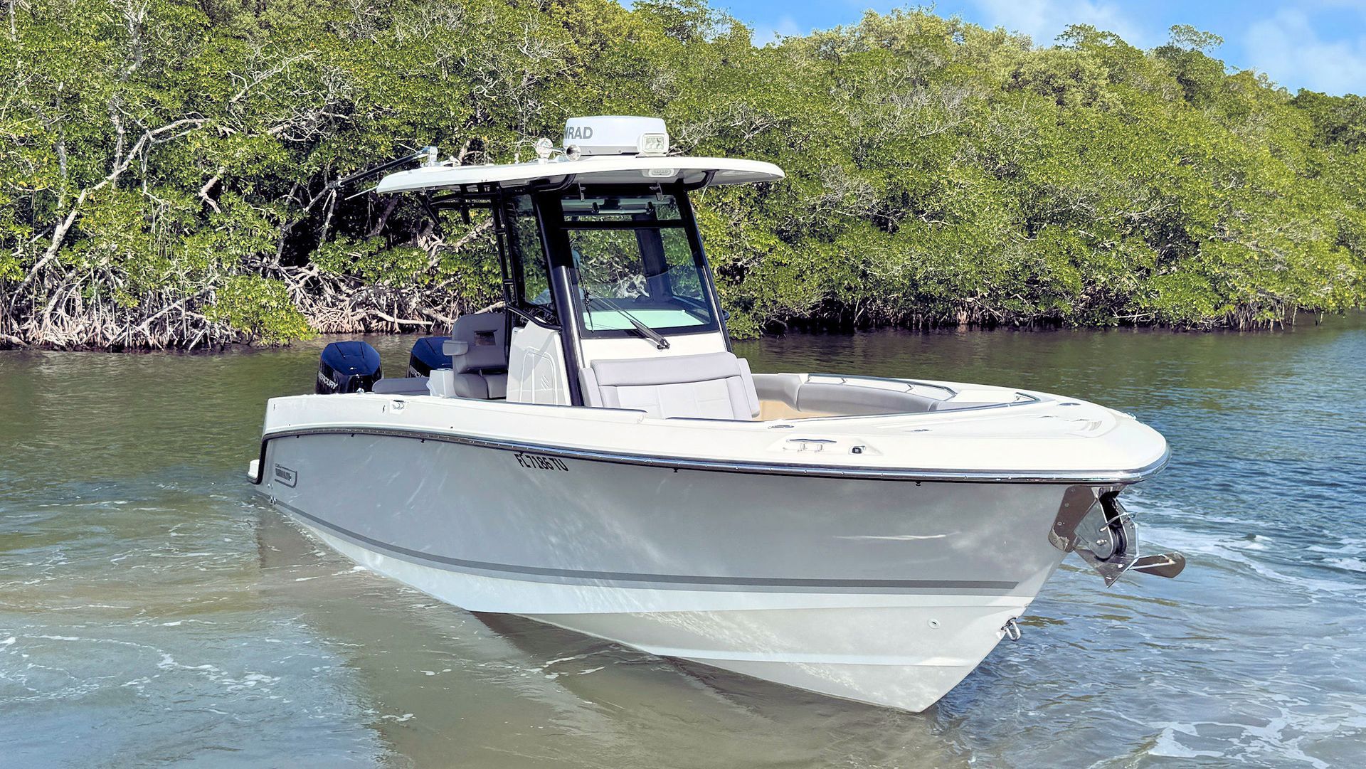 A white Boston Whaler center-console boat moving through calm water near a shoreline with dense green trees.