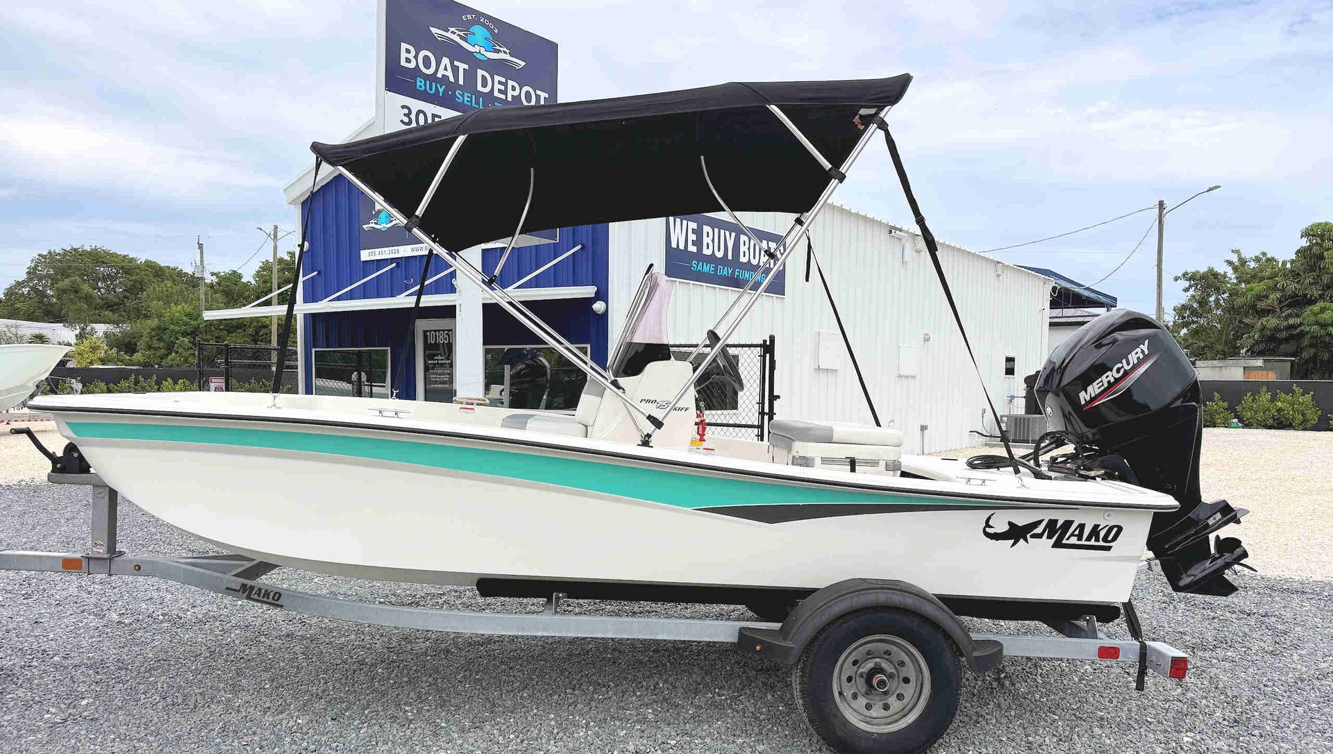 White and teal skiff on a trailer, with a black bimini top and outboard motor, parked on gravel.