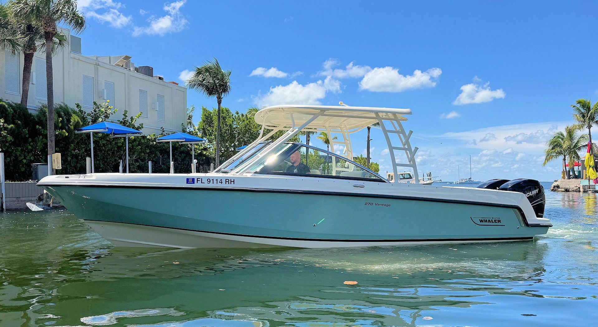 A light blue boat with a white  hard top travels through calm, sunny coastal waters near buildings and palm trees.