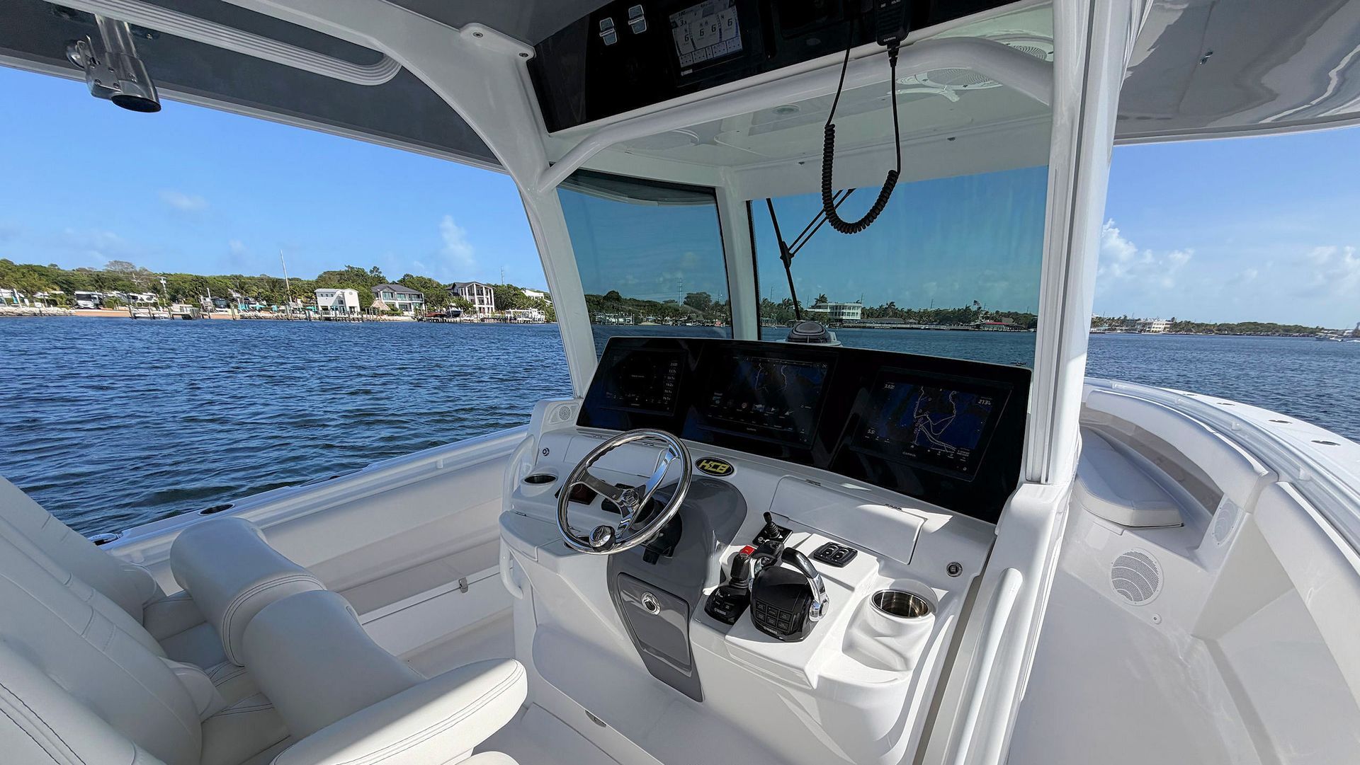 Boat helm with white seating and steering controls on a sunny day, overlooking blue water and shoreline