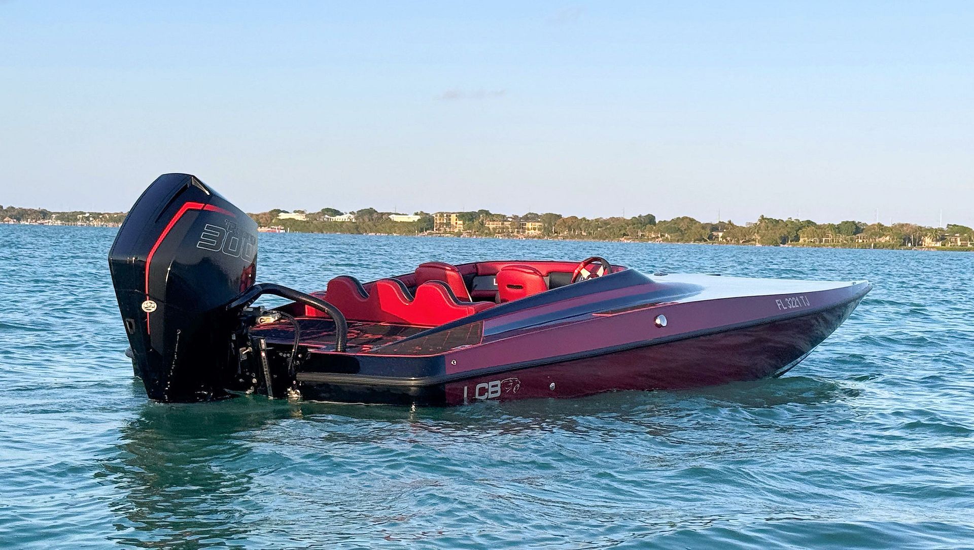 Red high-performance catamaran with a Mercury 300R outboard floating on blue water near a shoreline under a clear sky