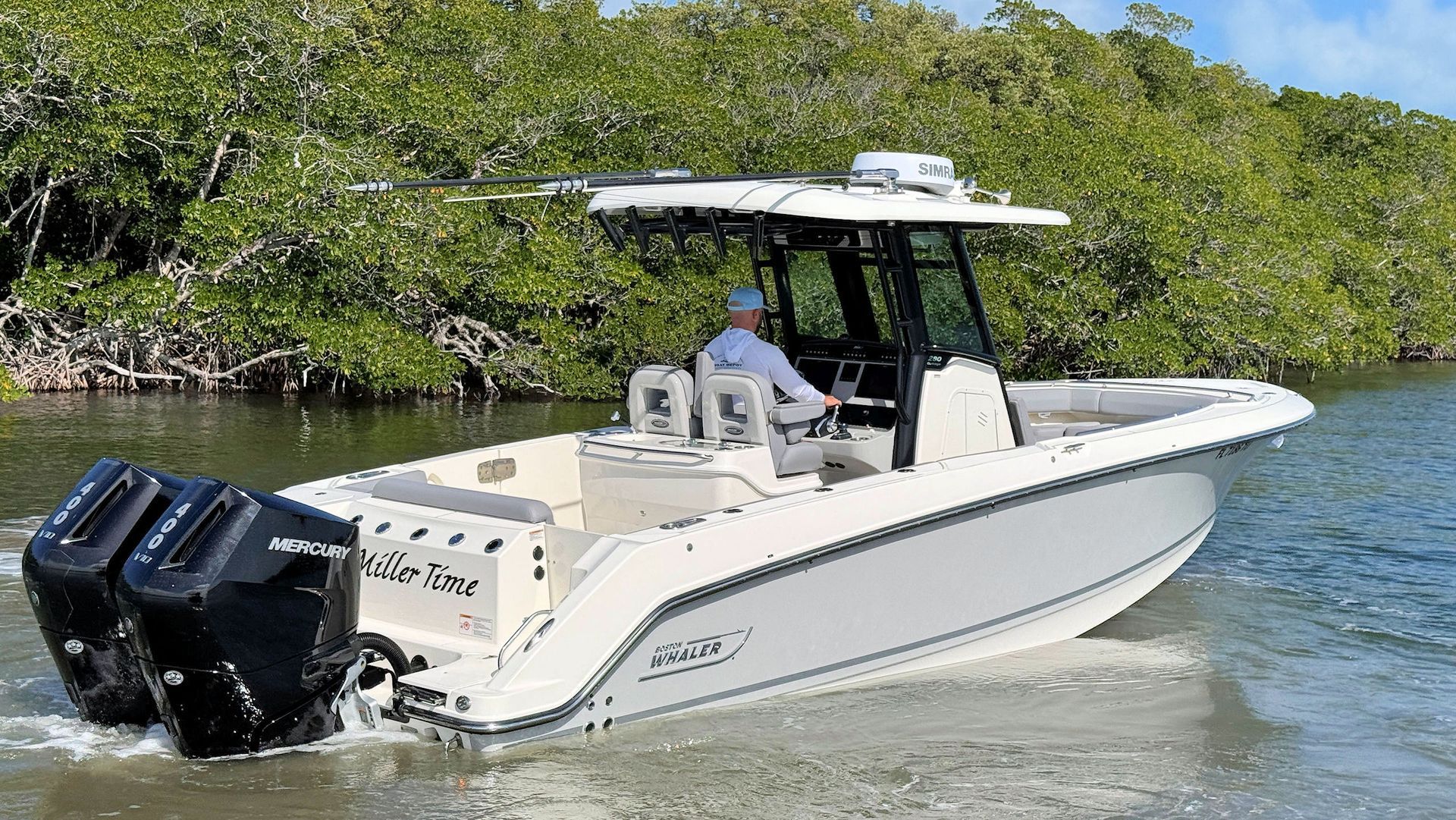 A white Boston Whaler center console boat with twin black outboard engines travels through shallow water near a wooded shoreline.