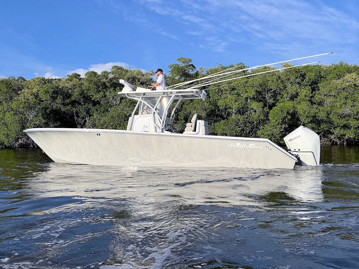 A white boat with a man on top, in water, near a line of green trees on a sunny day.