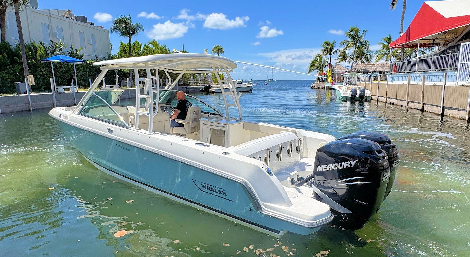 A light blue motorboat with a hardtop and two black outboard motors docked in a sunny, tropical marina.