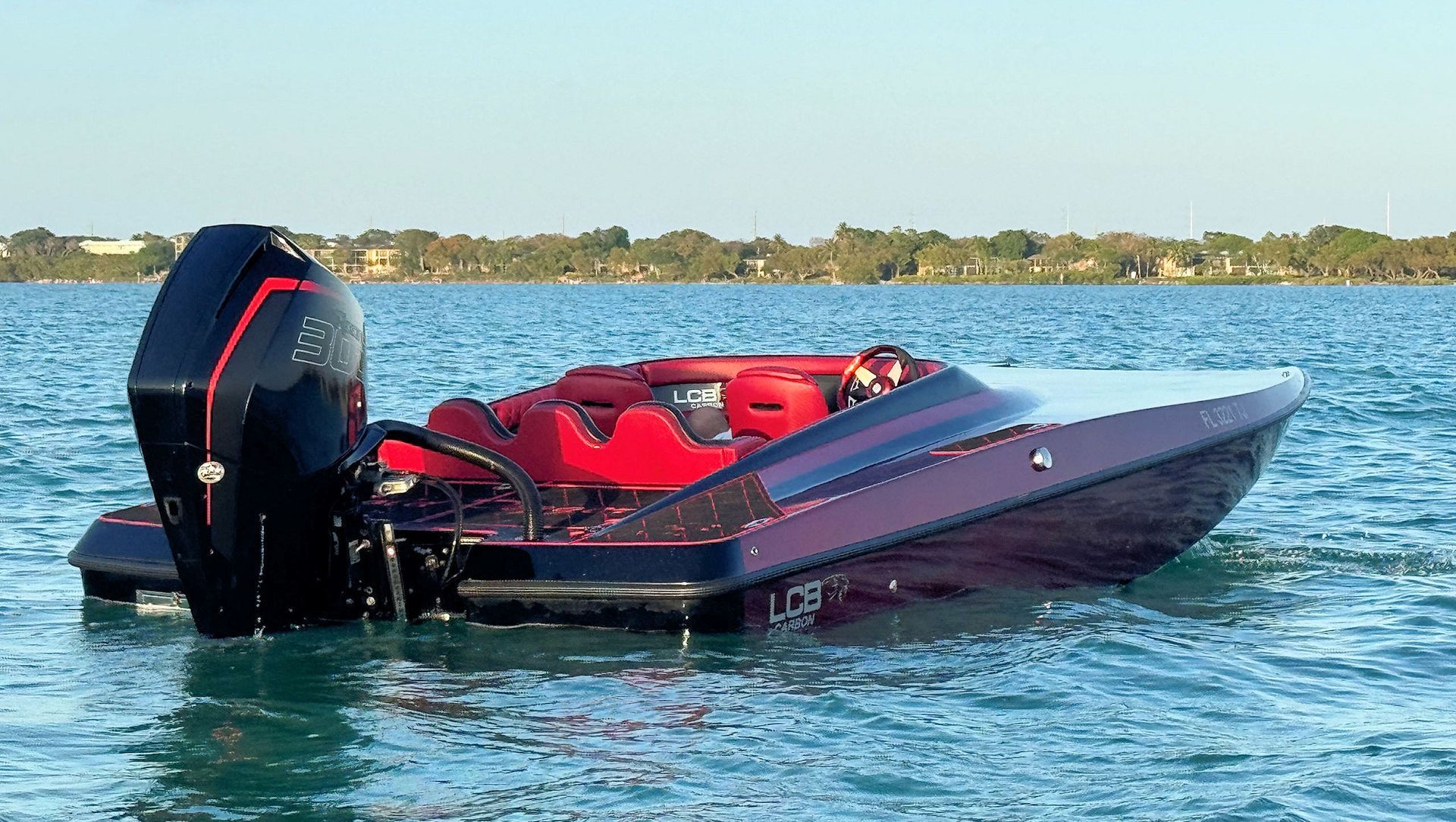 Red high-performance catamaran with black outboard motor floating on calm water near a distant shoreline.