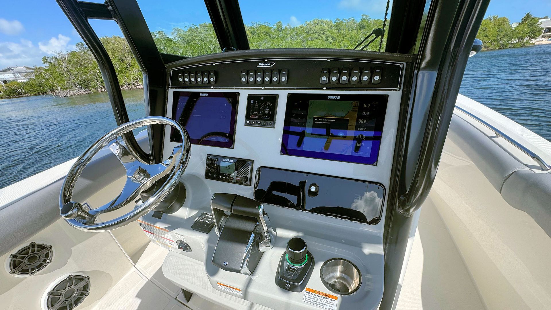 The console and steering wheel of a boat, featuring navigation screens and controls, overlooking a body of water.