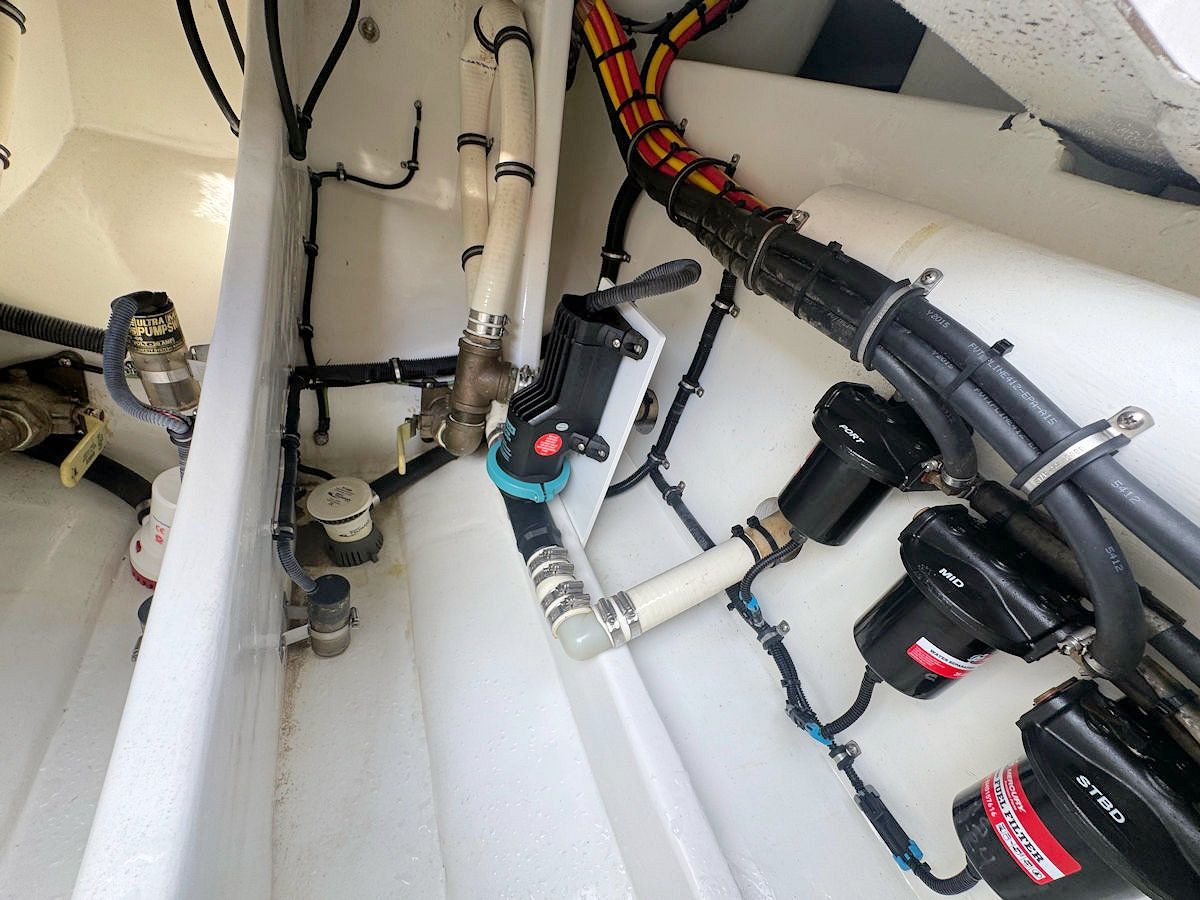 Boat's engine compartment: pipes, pumps, and wires against a white surface, with a black and red valve visible.