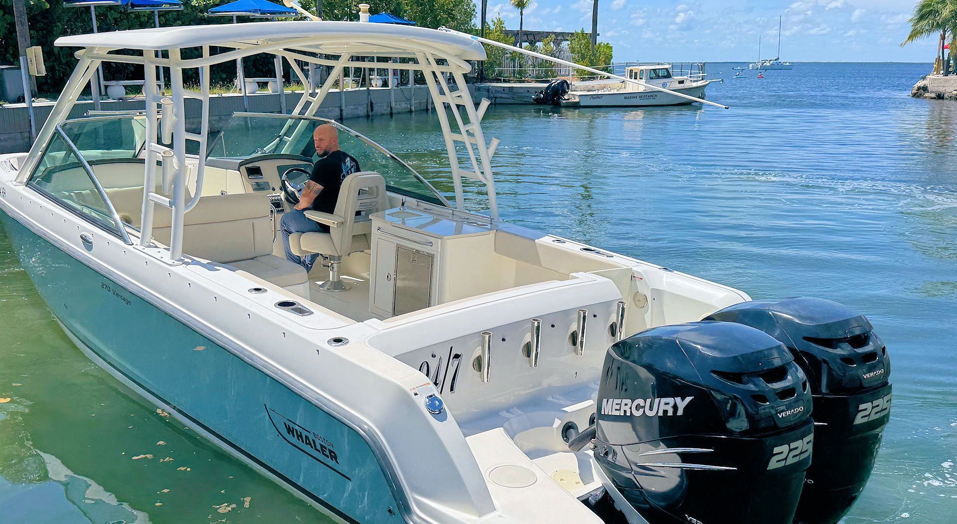 A person sits at the helm of a light blue and white motorboat with twin Mercury outboard motors at a sunny marina.