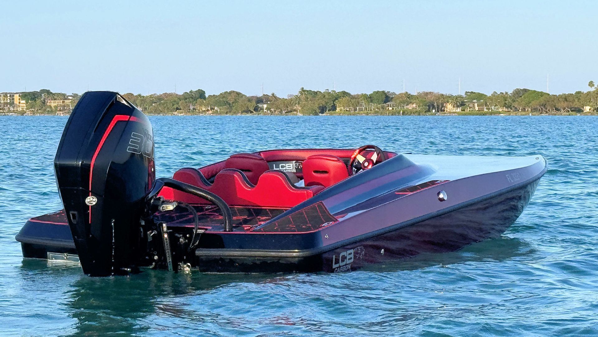 Red high-performance catamaran with black outboard motor floating on calm blue water near a shoreline