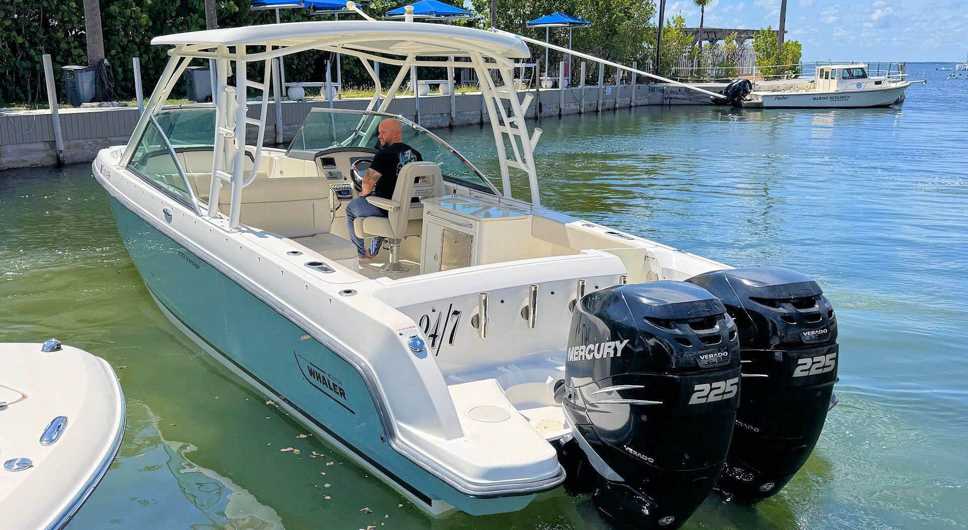 A light blue and white dual-outboard motorboat docked at a marina on a sunny day.