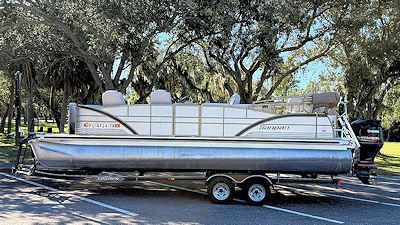 Tritoon boat on trailer parked on asphalt, near trees.