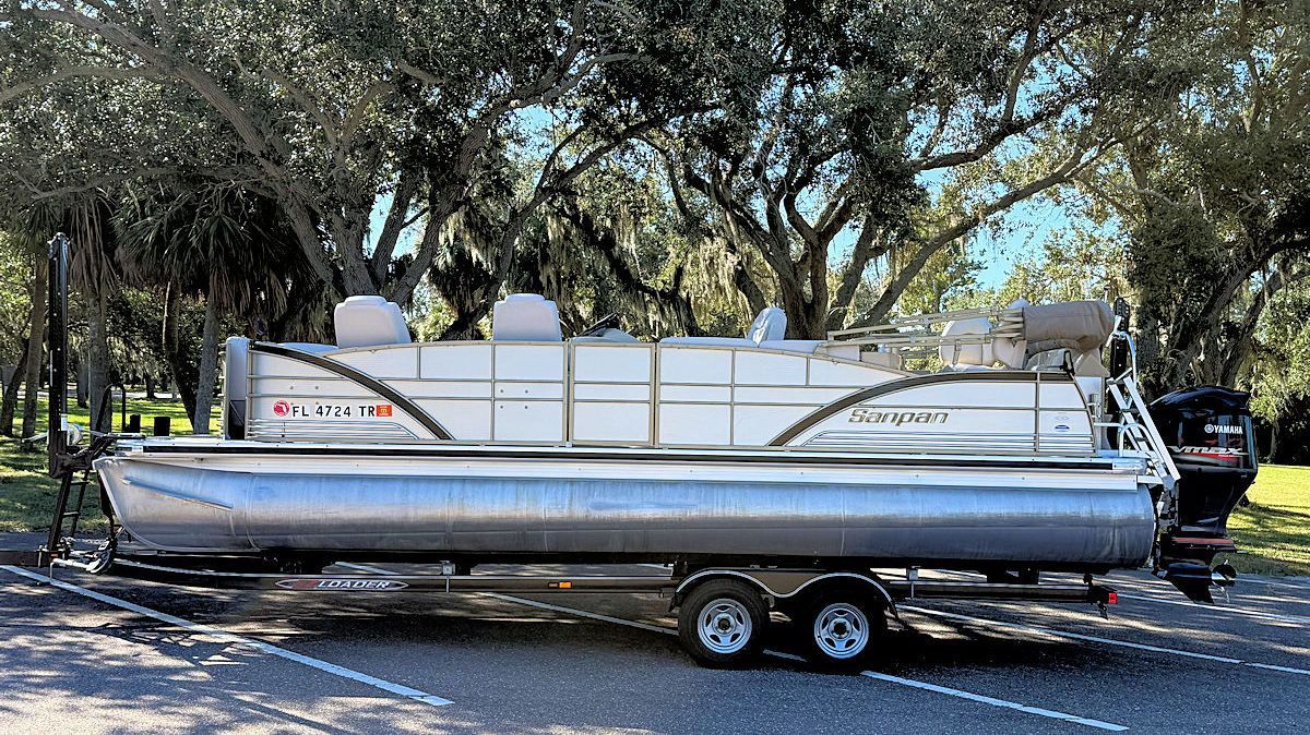 Pontoon boat on trailer, parked on pavement; trees in background.