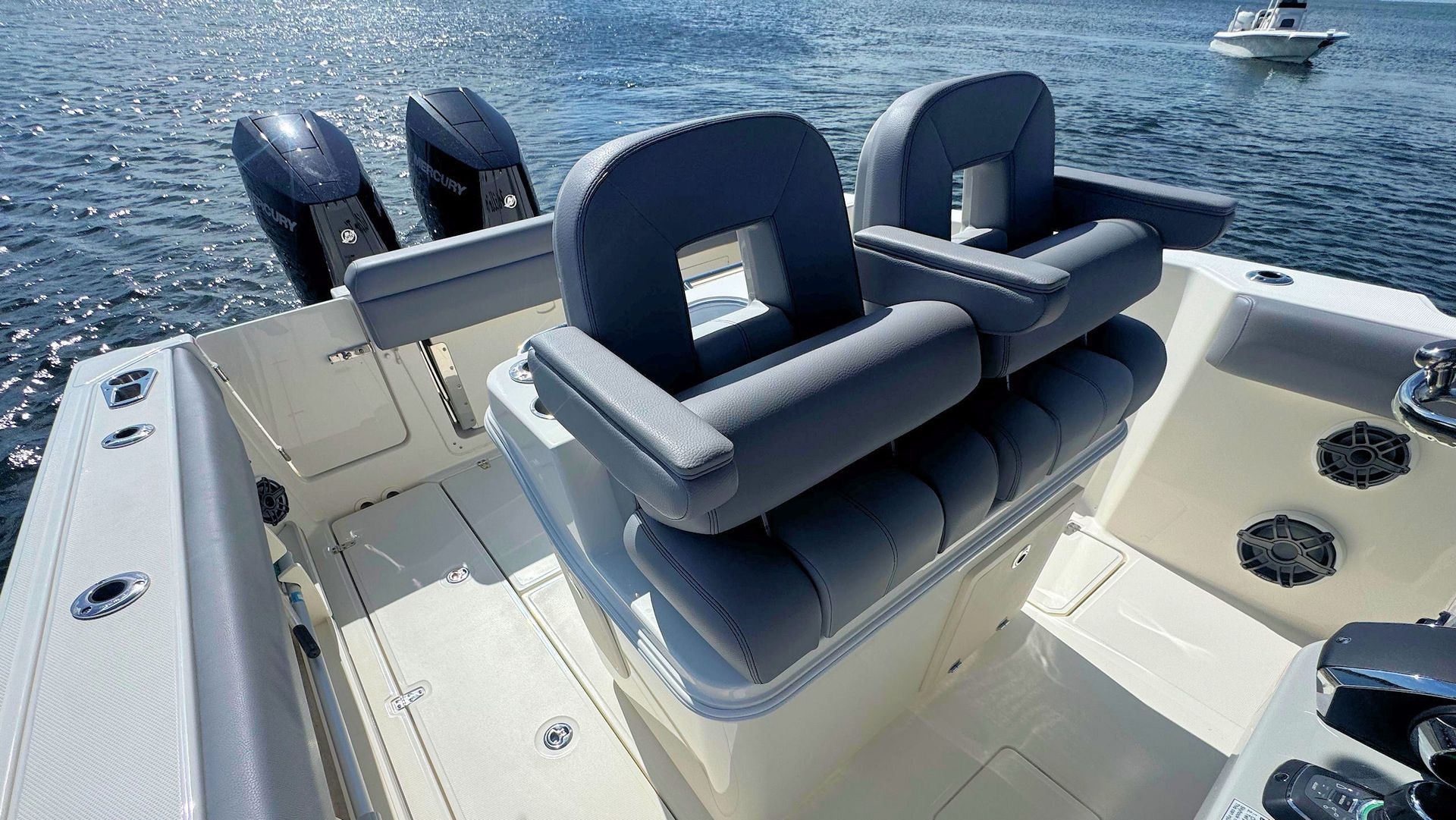 View from behind the helm of a boat, featuring two gray captains chairs with bolsters up, a cream interior, and two outboard motors.