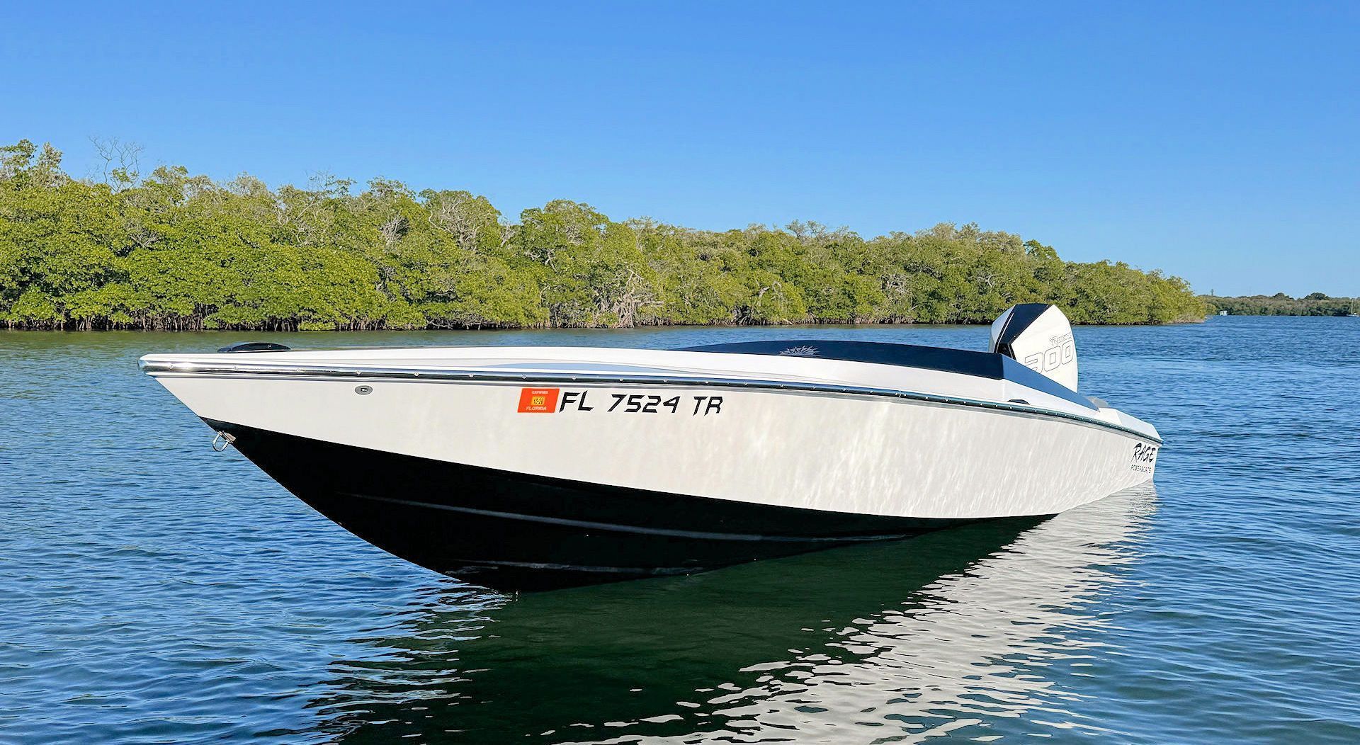 A white and black performance motorboat sits on calm blue water in front of a shoreline of green mangrove trees.