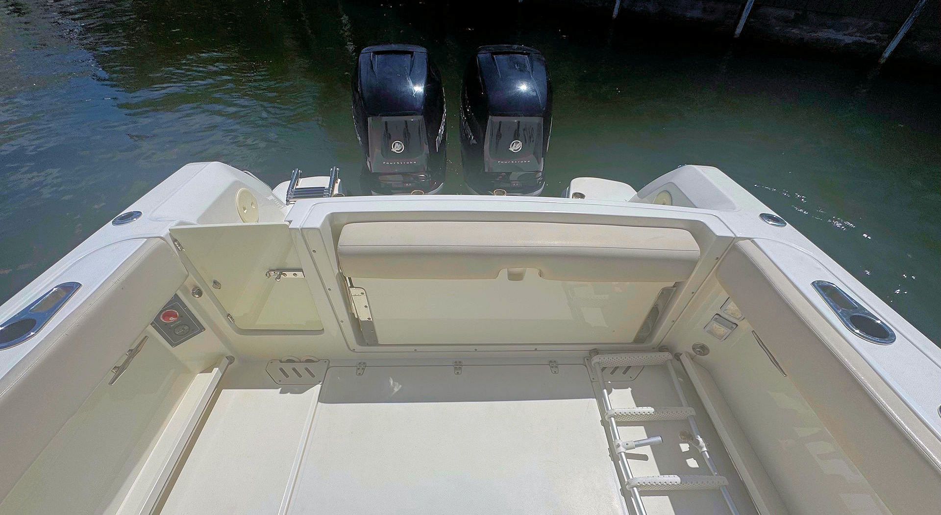 A high-angle view of a boat's white cockpit and aft deck, featuring a beige bench seat and two black outboard motors.
