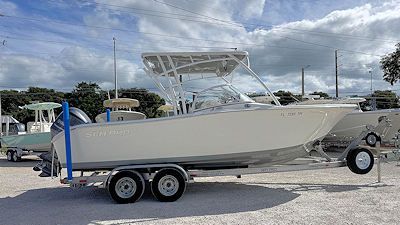 White boat on a trailer, parked on gravel. Blue sky, fluffy clouds, and another boat are visible.