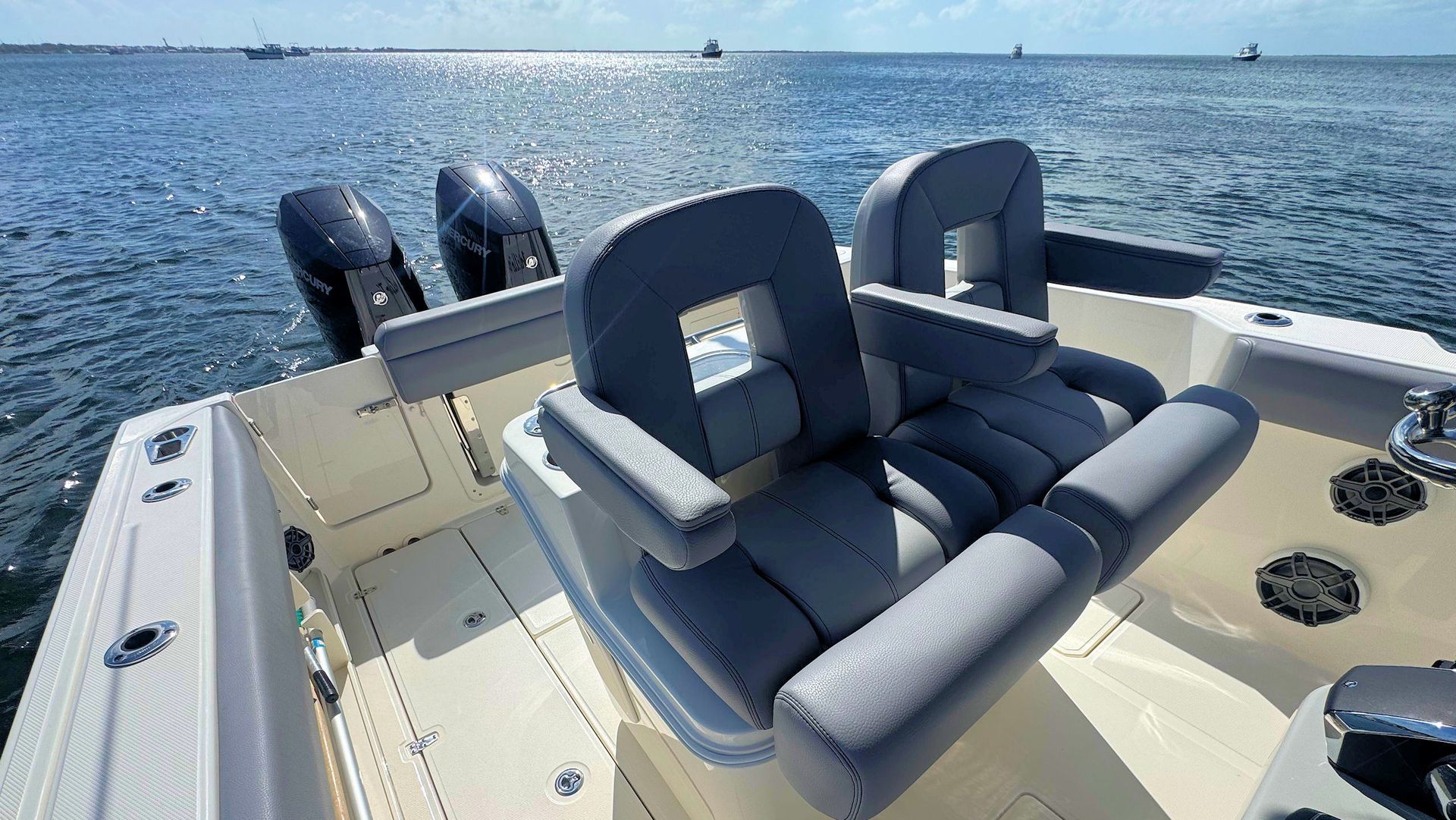 Two grey padded boat seats with armrests on a cream deck, facing outboard motors on a boat in sunny open water.