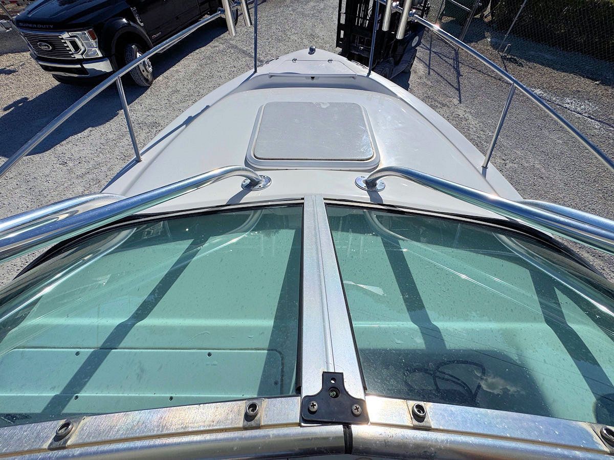 Boat's windshield, bow view. Tinted glass, metal frame, curved rails. Gray deck, sunny outdoor setting.