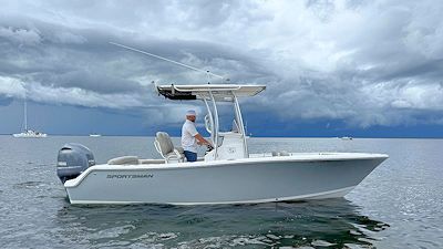 Man piloting a gray boat on calm water under a cloudy sky.