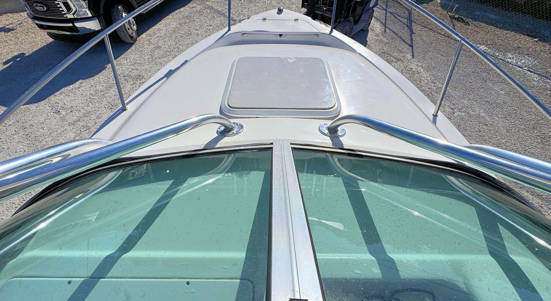 A view looking over the bow of a light-colored boat, featuring a glass windshield, metal railings, and a deck hatch.