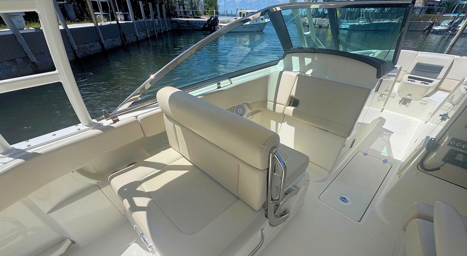 The interior of a boat cockpit with light beige upholstery, a clear windshield, and a view of the water and a dock.